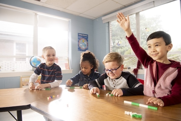 Four elementary students working together at a table in a classroom, with one student raising his hand.