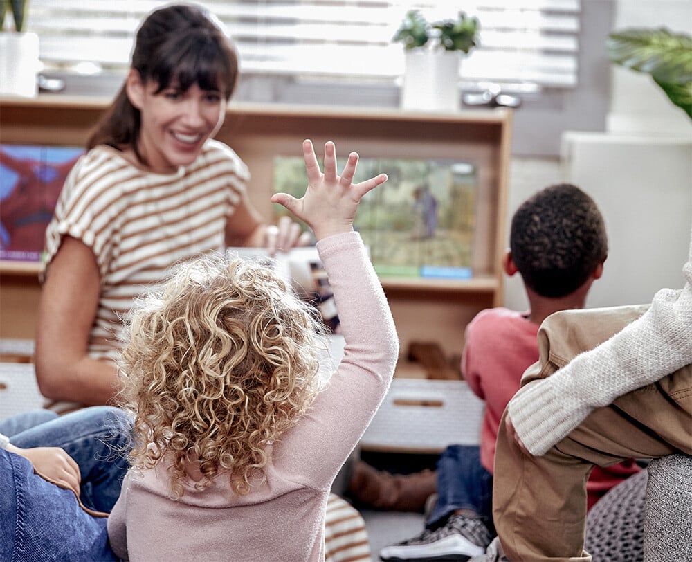 Student raising their hand as an educator reads them a Geodes book