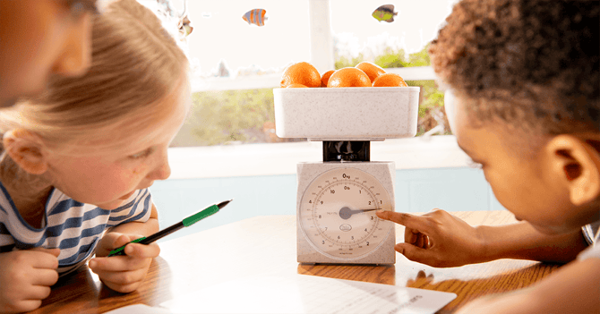 Three elementary students engage in a hands-on learning activity using a scale to weigh a bowl of oranges. One student points to the scale's dial while another takes notes with a pen, fostering collaboration and practical application of math concepts, as part of a Eureka Math² lesson.